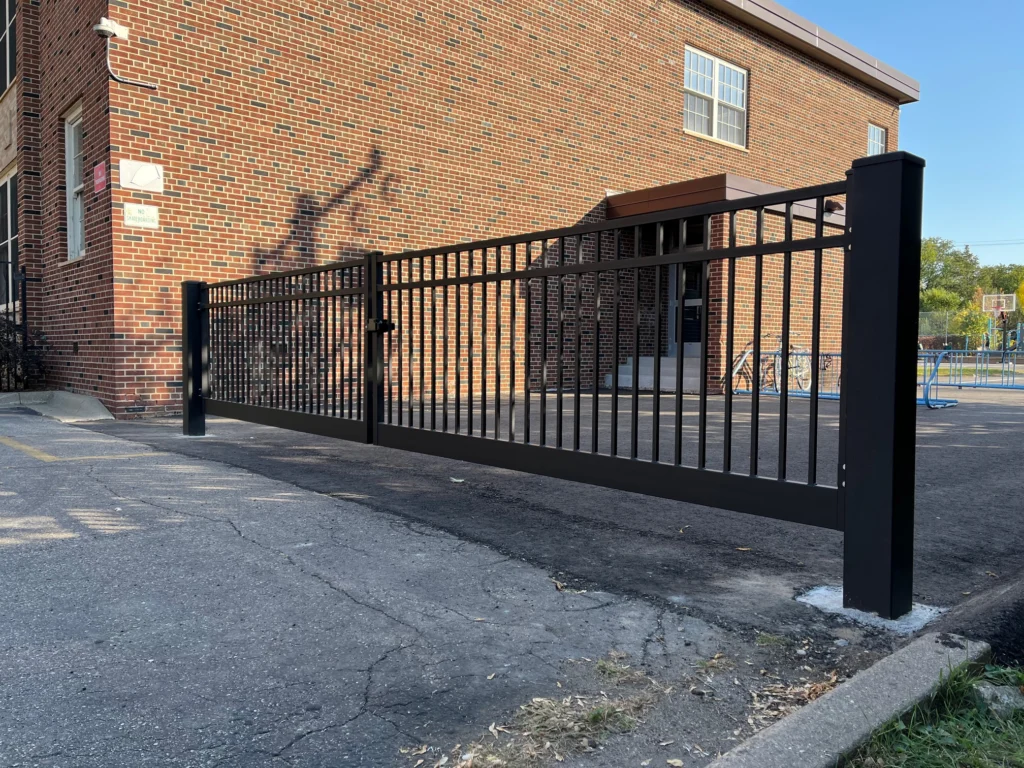 Black metal security fence with double gate installed along a paved driveway beside a brick commercial building.