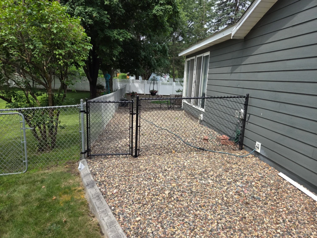 Black chain link fence with gate enclosing a residential side yard with gravel landscaping next to a house.