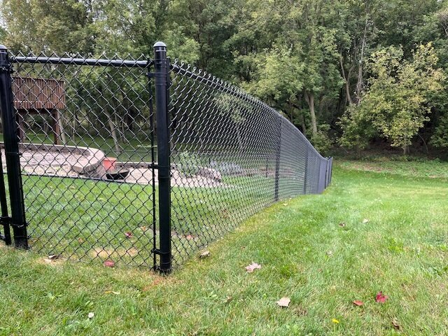 Chain link fence enclosing a large open field or sports area under a blue sky with scattered clouds.