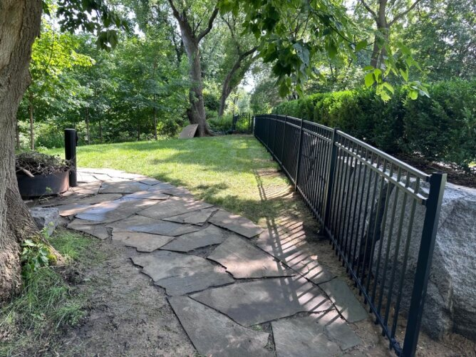 Black metal fence running alongside a landscaped garden path with trees, grass, and natural stone walkway.