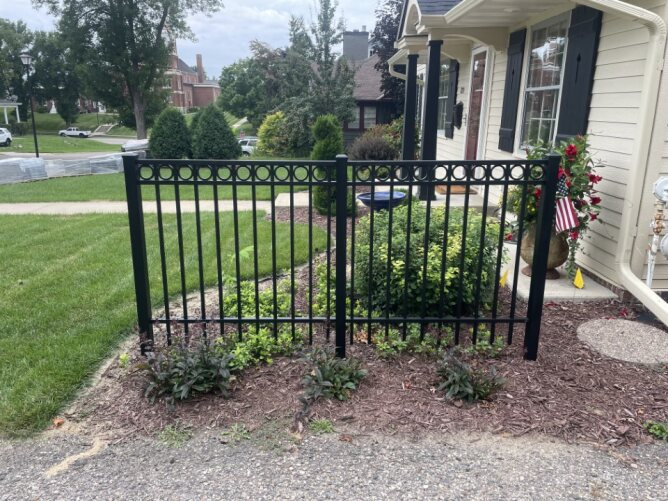 Black metal ornamental fence with a curved gate installed in a landscaped residential yard with grass and trees.