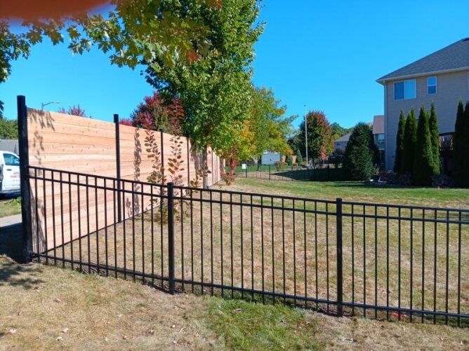. Black metal fence surrounding a landscaped residential yard with trees, shrubs, and lawn area.