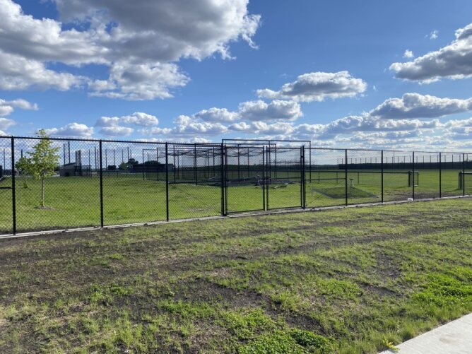 Chain link fence enclosing a large open field or sports area under a blue sky with scattered clouds.