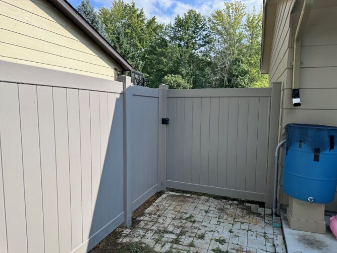 Gray vinyl privacy fence with gate installed between two houses, enclosing a small patio area with paver flooring.