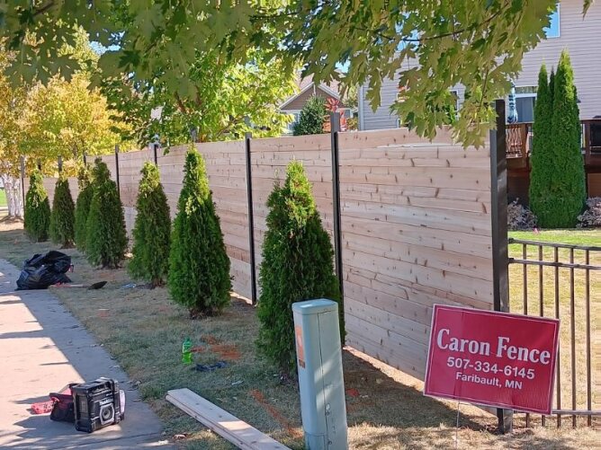 Horizontal wooden privacy fence with black metal posts installed along a residential sidewalk with trees and landscaping.