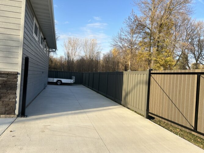 Modern composite privacy fence with gate installed along a driveway beside a house with trees and a parked trailer.