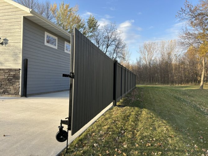 Modern dark composite privacy fence with gate installed along a driveway beside a house and open grassy area.