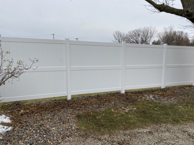 White vinyl privacy fence installed along a residential yard with gravel and grass ground.