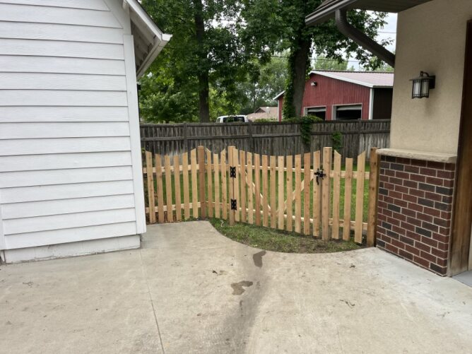Wooden picket fence with double gate installed near a house patio, enclosing a backyard area.