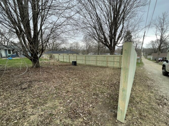 Wooden privacy fence enclosing a residential yard with trees and dirt ground along a gravel driveway.