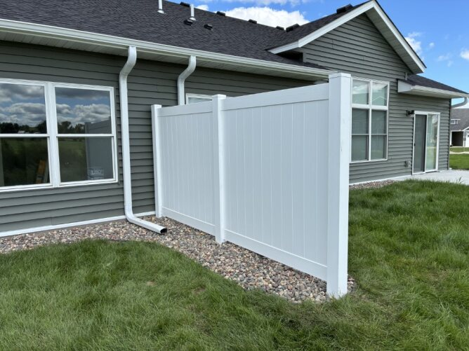 White vinyl privacy fence installed along the side of a residential house with green lawn and drainage system.