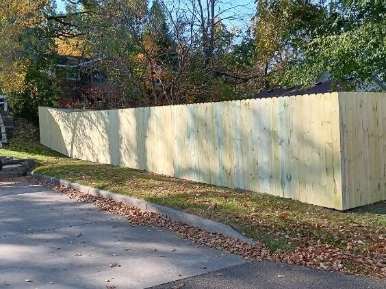 Long wooden privacy fence installed along a residential street with trees and fallen leaves in the background.