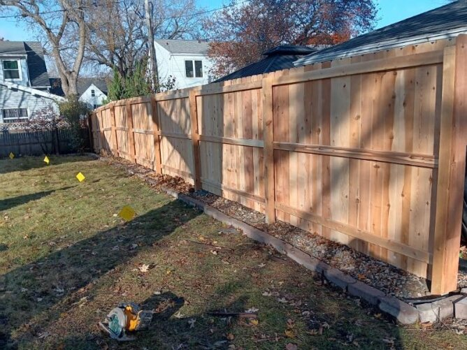 Newly installed wooden privacy fence along a residential property with dirt ground and trees nearby.