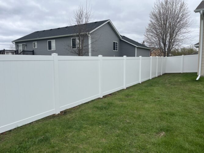 White vinyl privacy fence installed along the side of a house beside a lawn and parked trailer.