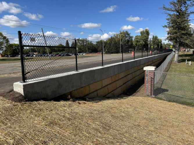 Black chain link fence installed along a sidewalk enclosing a large open field and parking area.