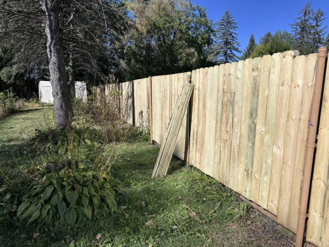 Long wooden privacy fence installed along a residential street with trees and fallen leaves in the background.