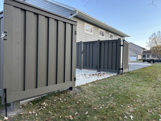 Modern dark composite privacy fence with gate installed along a driveway beside a house and open grassy area.