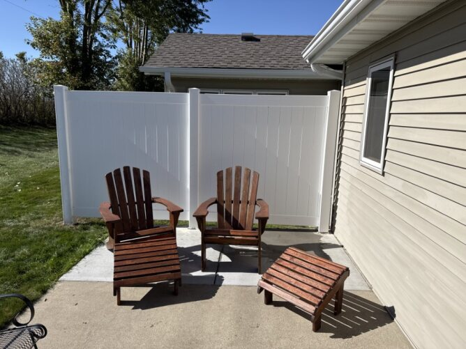 White vinyl privacy fence enclosing a backyard patio with wooden outdoor chairs and a small table.
