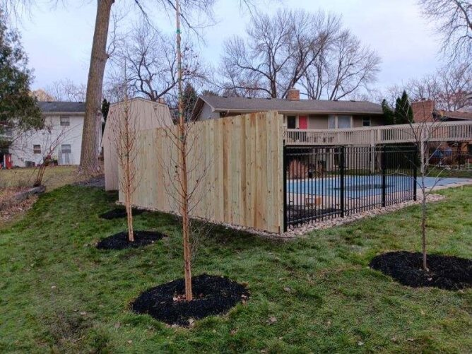 Horizontal wooden privacy fence with black metal posts installed along a residential sidewalk with trees and landscaping.