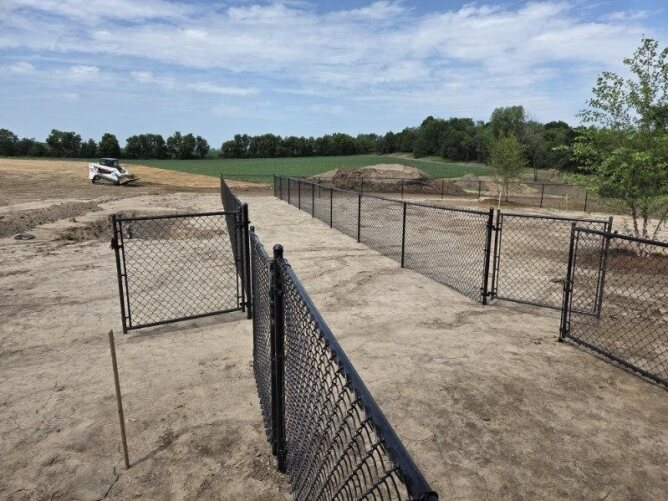 Black chain link fence installed along a sidewalk enclosing a large open field and parking area.