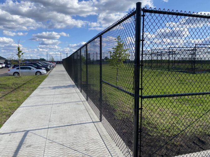 Black chain link fence installed along a sidewalk enclosing a large open field and parking area.